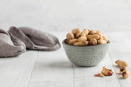 Peanuts in a shell in a gray bowl on a white wooden background. Healthy snacks, vegetable fats. Copy...