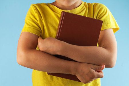 A schoolboy child in a yellow t-shirt holds a book on a blue background. The concept of the beginning of training, September 1, teacher's day.の写真素材