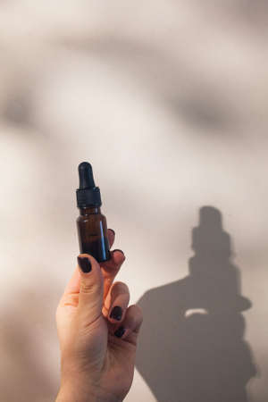 A womans hand holds a bottle of face oil with hard shadows on the background. Concept of beauty.