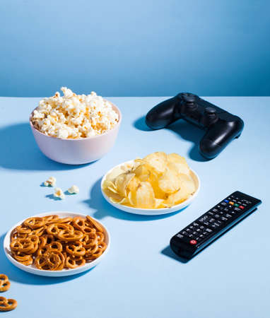 Assortment of unhealthy snacks, such as chips, popcorn, pretzels and a TV remote, a game joystick on a blue background.の写真素材