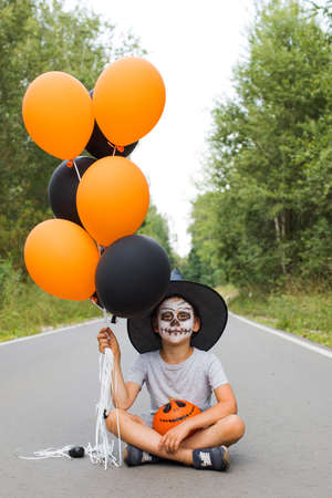 Boy with skeleton makeup and hat holds pumpkin and balloonsの写真素材