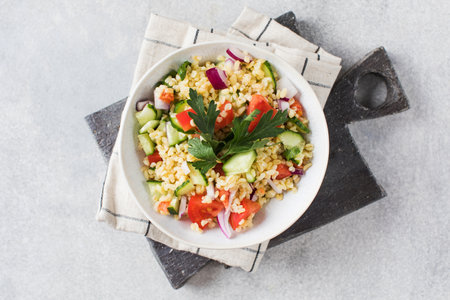 Vegetable and bulgur salad in deep bowl on white table decorated with board and towelの写真素材