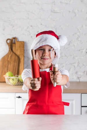 Cute little boy in Santa hat and apron shows rolling pin and whisk in kitchenの写真素材