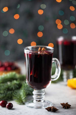 A glass of hot spicy mulled wine surrounded by spices and branches of a Christmas tree with bokeh in...