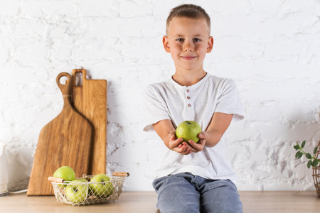 A cute blond boy is holding a ripe green apple in his hands in the kitchen.の写真素材