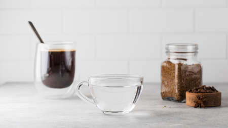 On the grey table there is a mug of water, a mug of black coffee and a jar of instant coffee.