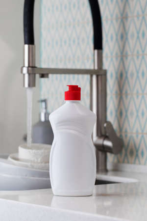 A white bottle with dishwashing gel on the background of a sink with dirty dishes and pouring water...