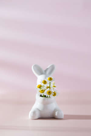 Figurine of a white rabbit with a bouquet of white daisies on a pink background with a shadow pattern. Happy Easter.の写真素材