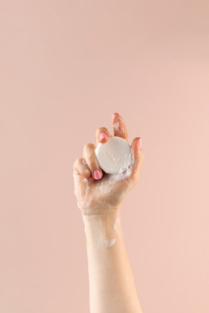 A womans hand holds a round piece of soap on a light pink background.