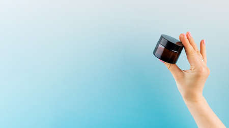 Mockup of a glass brown jar with a cosmetic product in a female hand on a blue background. Copy space.の写真素材