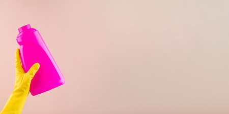Hand in protective glove holds bright pink bottle of detergent on beige closeup