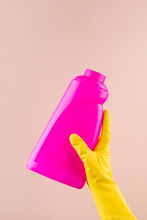 Hand in protective glove holds bright pink bottle of detergent on beige closeupの写真素材
