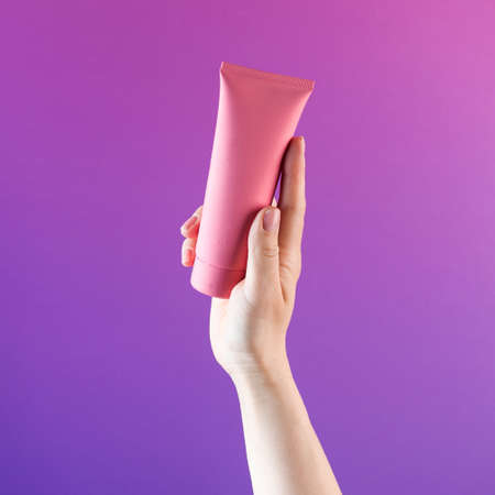 Mockup of a pink tube with a cosmetic product in a female hand on a purple background. Copy space. Banner.の写真素材