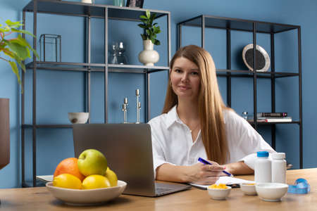 A female nutritionist smiles at the camera, sitting at her desk in a white shirt at a laptop.の写真素材