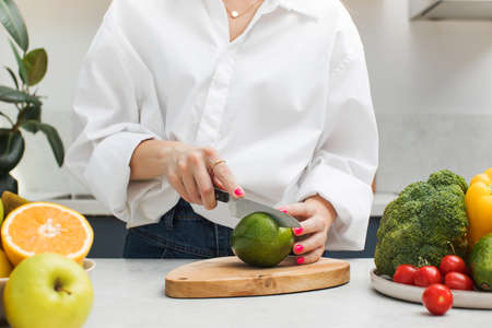 A young woman cuts a ripe avocado in half on a board on the kitchen table. Vegetarianism, healthy nutrition.の写真素材