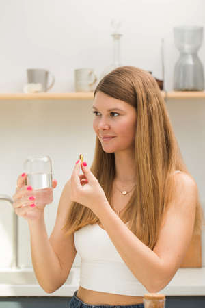 Happy blonde woman holding a glass of water and a capsule of fish oil or vitamin D in the kitchen....