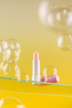 Pink moisturizing hygienic lipstick on a glass shelf on a yellow background surrounded by soap bubbles.の写真素材