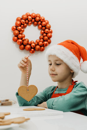 A cute boy in a Santa hat holds ginger cookies on a rope while sitting in the kitchen.の写真素材