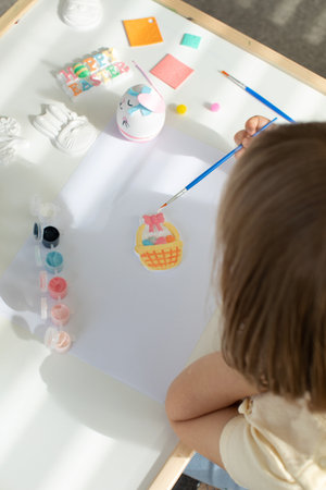 A cute little girl is sitting at a table and doing Easter crafts, coloring Easter figurines....