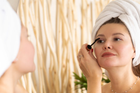 An attractive young woman paints her eyelashes with mascara in front of the bathroom mirror.