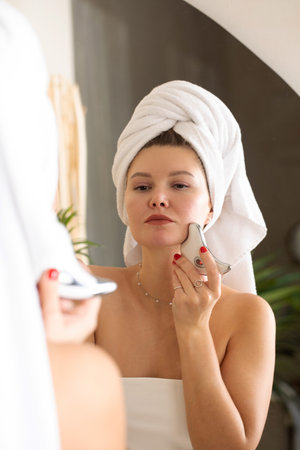 An attractive woman does a facial massage with a microcurrent massager in front of a bathroom mirror.の写真素材