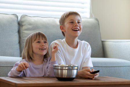 Cute joyful brother and sister are sitting at the table with a bowl of popcorn and watching TV. childrens leisureの写真素材