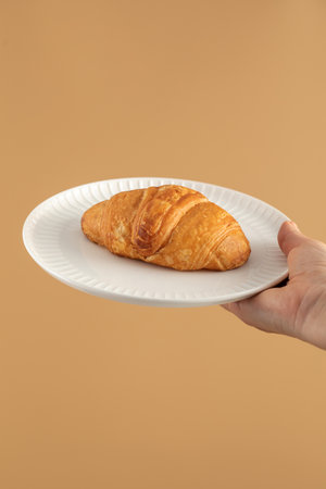 A womans hand holds a plate with a croissant on a brown background. Healthy breakfast.の写真素材