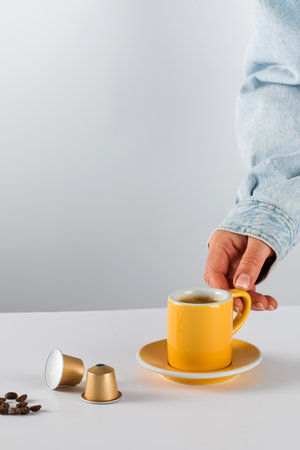 Hand reaching for a yellow coffee cup with golden coffee capsules and beans on a white backgroundの写真素材