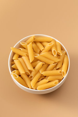 Uncooked penne pasta in a bowl on a beige background, Italian cuisine.の写真素材