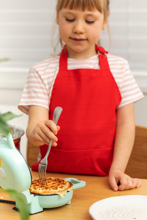 Junior schoolgirl chef checks waffle on mini oven with forkの写真素材