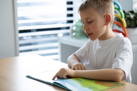 The boy is enthusiastically reading a book at a table in a bright room. Childrens leisure