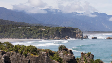 Scenic coastal landscape with rock formations and mountain backdrop. Punakaiki Pancake Rocks, New...