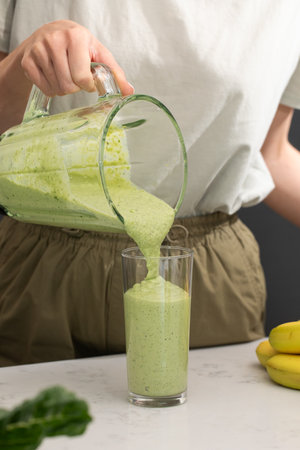 Woman pouring green smoothie from blender into glass in kitchen. Detox, vegan food