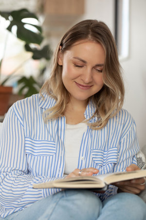 Smiling young woman reads bestseller book sitting in living roomの写真素材