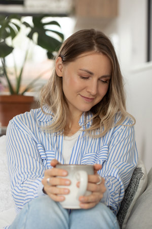 Pretty woman holds tea mug sitting on comfortable sofaの写真素材