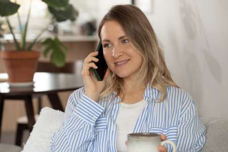 Woman with coffee cup talks on cellphone sitting on sofaの写真素材