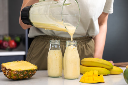 Woman pouring refreshing tropical smoothie with banana, pineapple and mango from blender into bottleの写真素材