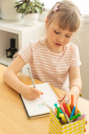 Cute little girl draws picture on notepad page at tableの写真素材