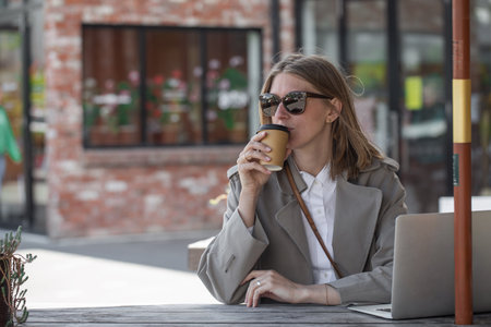 Caucasian businesswoman drinks coffee and works on laptop, sitting at table outdoor. Remote workの写真素材