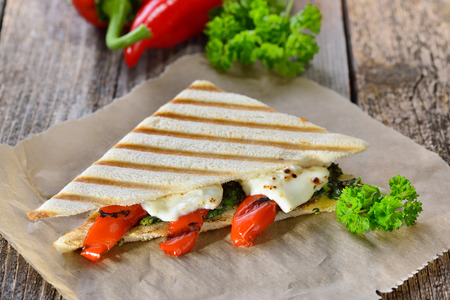 Vegetarian toast: Panini with grilled red bell peppers and feta cheese on pesto sauce, served on sandwich paper on a wooden tableの写真素材