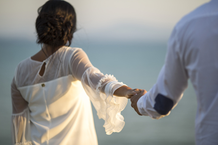 man and woman walking together grasping hands against a beautiful view of sea, ocean and marvelous sky. brides go to the shore of the seaの写真素材
