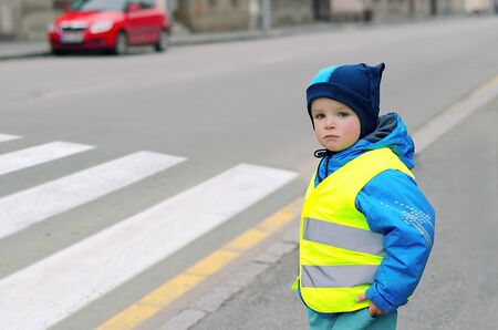 Child in front of pedestrian crossing. A little boy finds out if he can cross the crossswalk. He wears reflective vest because of safety. Car in the background. Child concept. Traffic concept.の写真素材