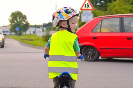 Little boy is going to cross road. . Boy wearing yellow reflective vest and helmet because of safety. There is a red car in the backgroundの写真素材