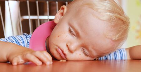 Cute blond boy falling asleep on the table. He is prepared for a eating : wearing a bib and sitting on the chair.の写真素材