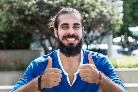 Man with beard and blue shirt showing both thumbs outdoor in the city with modern buildings in the backgroundの写真素材