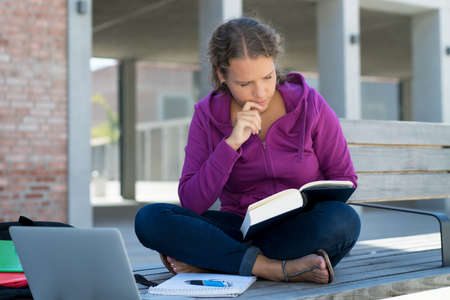 Beautiful german female student learning with book and computer in front of school building