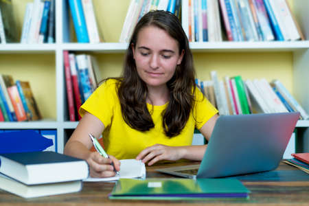 Pretty german female student learning with book and computer at deskの写真素材
