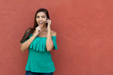 Latin american young adult woman with long dark hair talking at mobile phone with wall as copy space outdoor in summerの写真素材