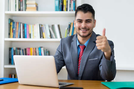 Laughing mexican businessman at computer showing thumb up indoors at officeの写真素材