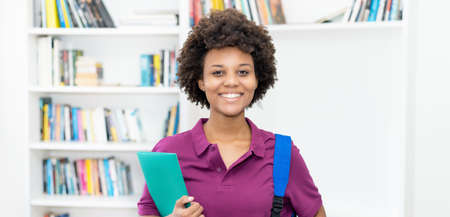 Laughing afro american female student with backpack and paperwork at classroom of universityの写真素材